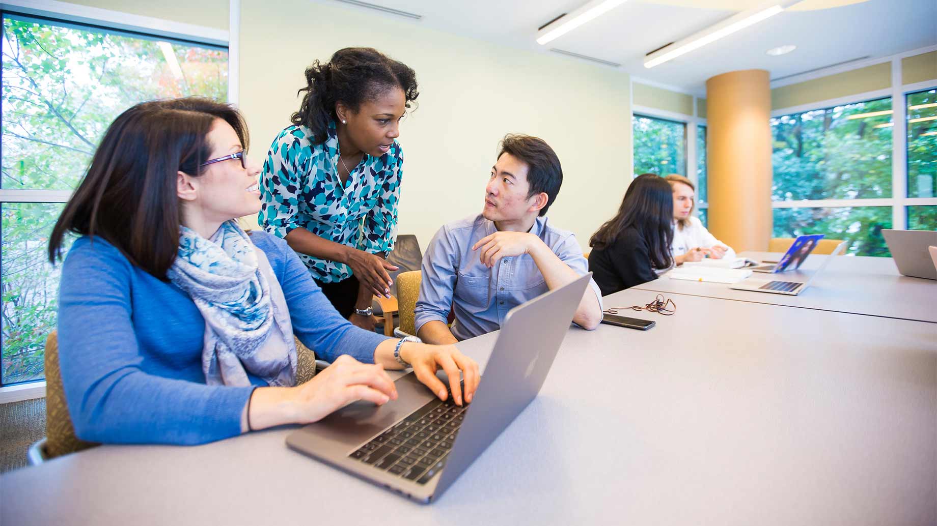 students sitting at a conference table with laptops and chatting with an instructor