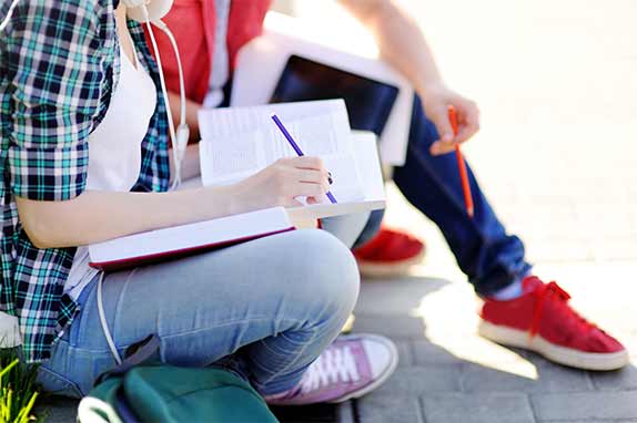 girl in plaid shirt writing in notebook with boy in red shirt and sneakers beside her