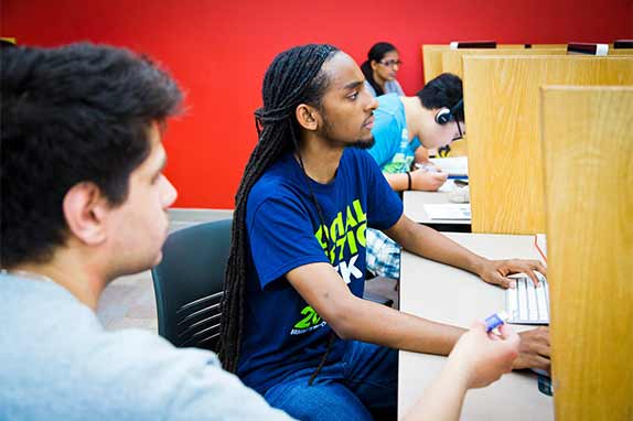 Young man with bright blue and green shirt with long dreadlocks at computer against a red backdrop