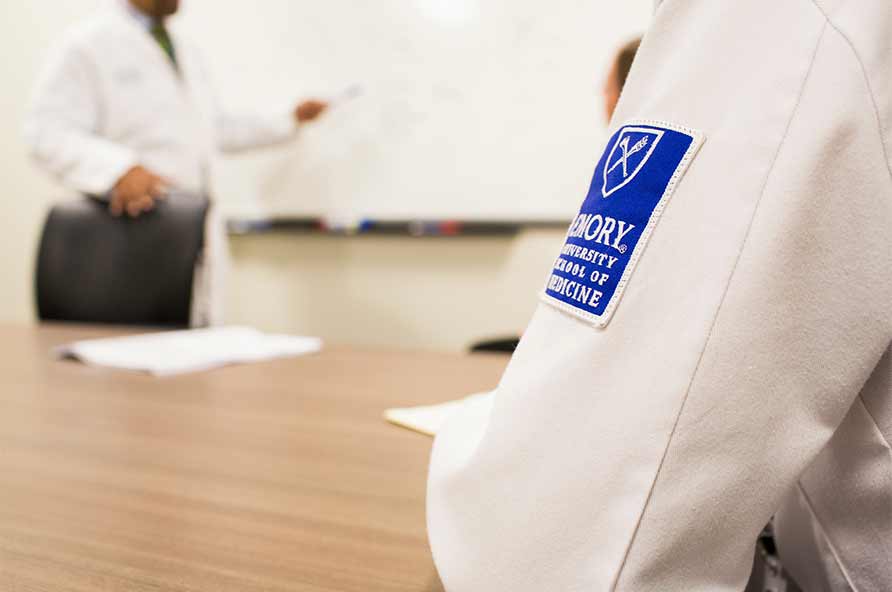 closeup of a white coat with an Emory School of Medicine patch on the sleeve and an instructor at a whiteboard in the background